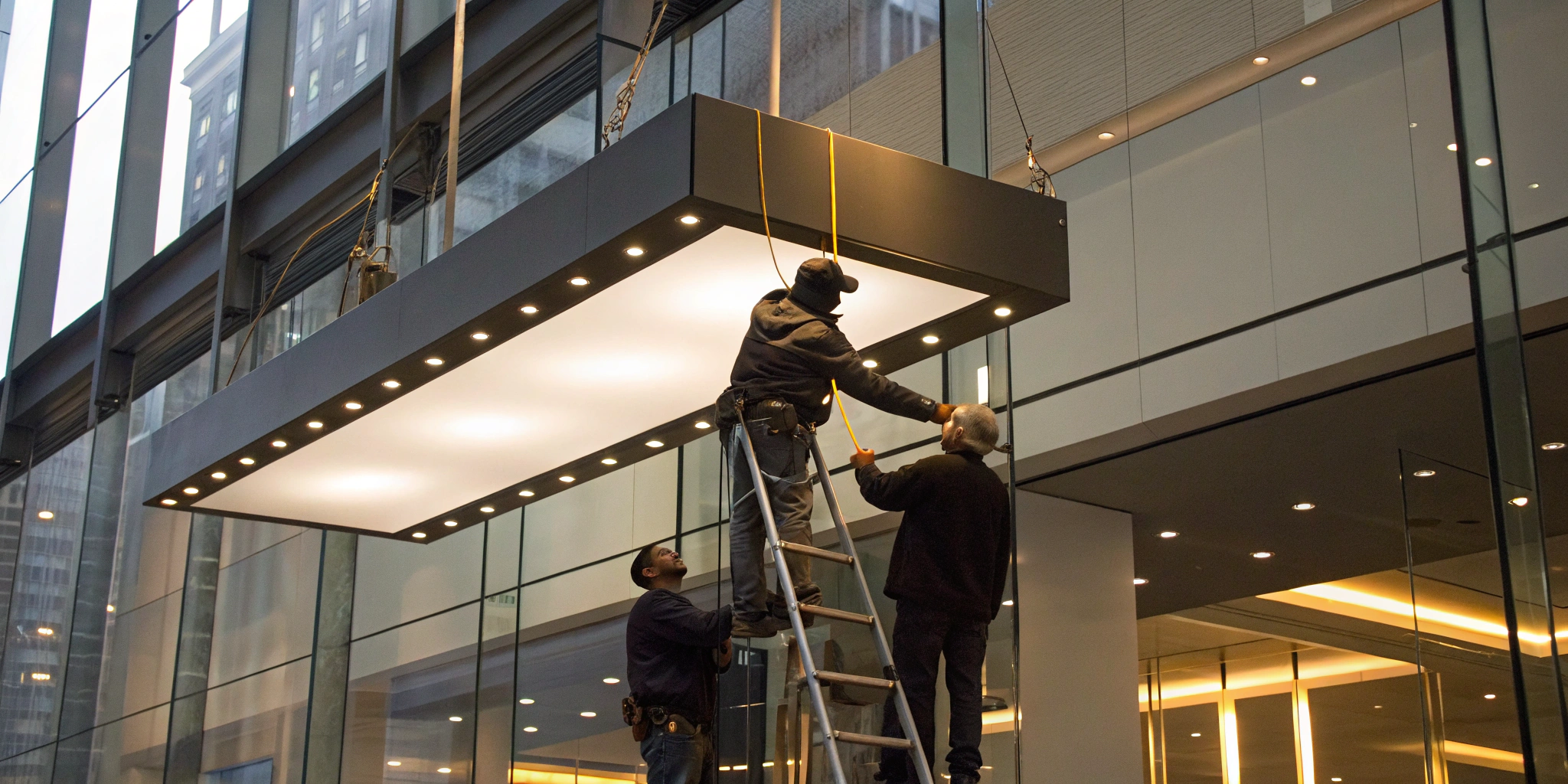 Workers installing a large illuminated hanging sign in a commercial building interior.